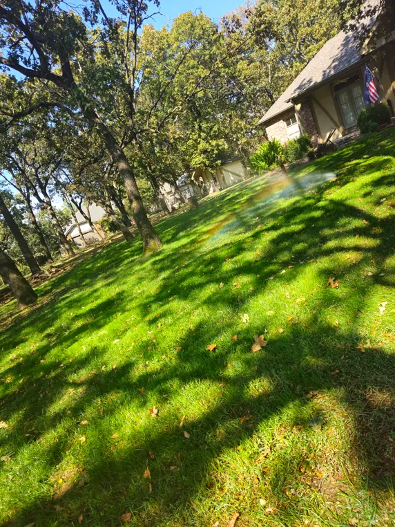 A sprinkler fan throwing water across a sloped Tulsa lawn under mature oaks, a faint rainbow standing in the mist